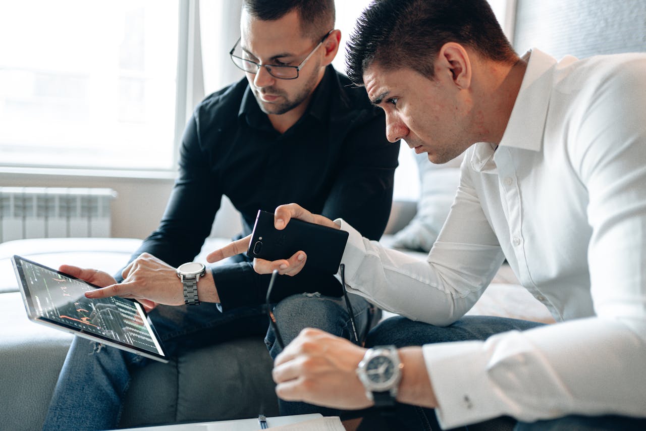 journey Two men analyzing financial data on a digital tablet indoors in a professional setting.